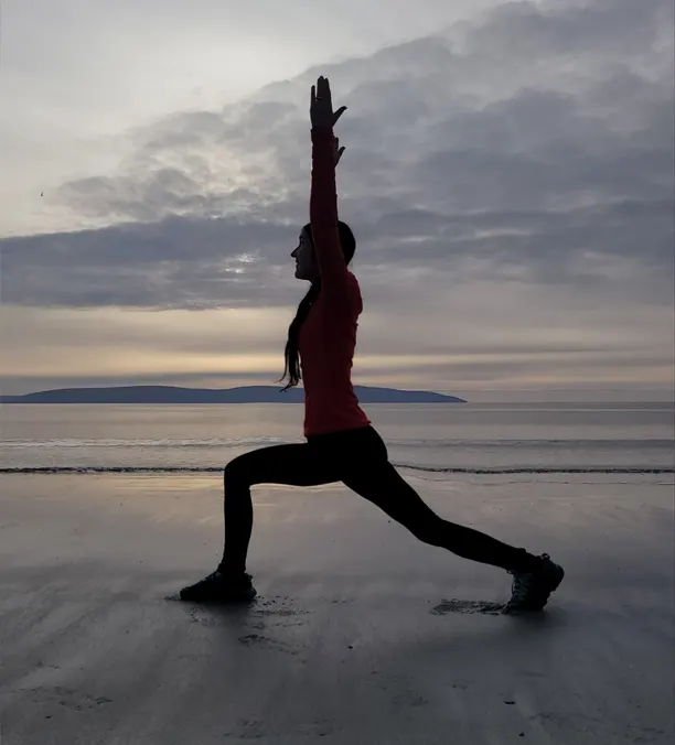 Mujer practicando yoga en la playa al amanecer, reflejando Yoga como Estilo de Vida.